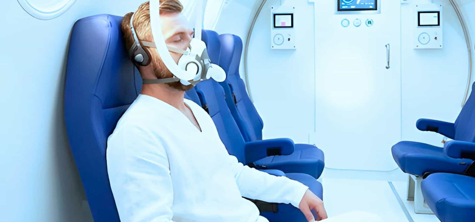 Man sitting in a clinical chamber wearing a breathing mask connected to medical equipment, undergoing a controlled respiratory or oxygen therapy test.