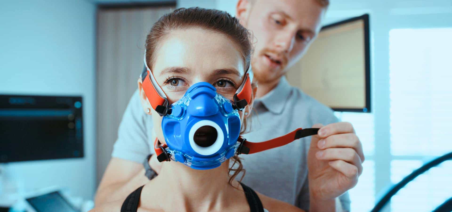 Woman wearing a respiratory training mask while a trainer adjusts the straps in a fitness or testing environment.