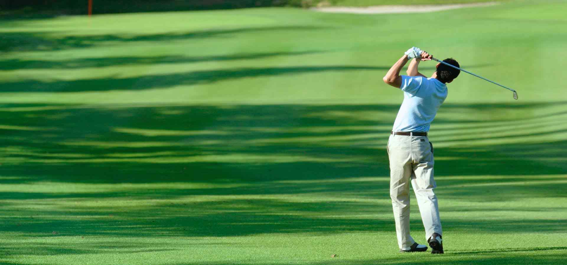 Golfer completing a swing on a well-maintained golf course, standing on a lush green fairway surrounded by rolling grass and soft shadows on a sunny day.