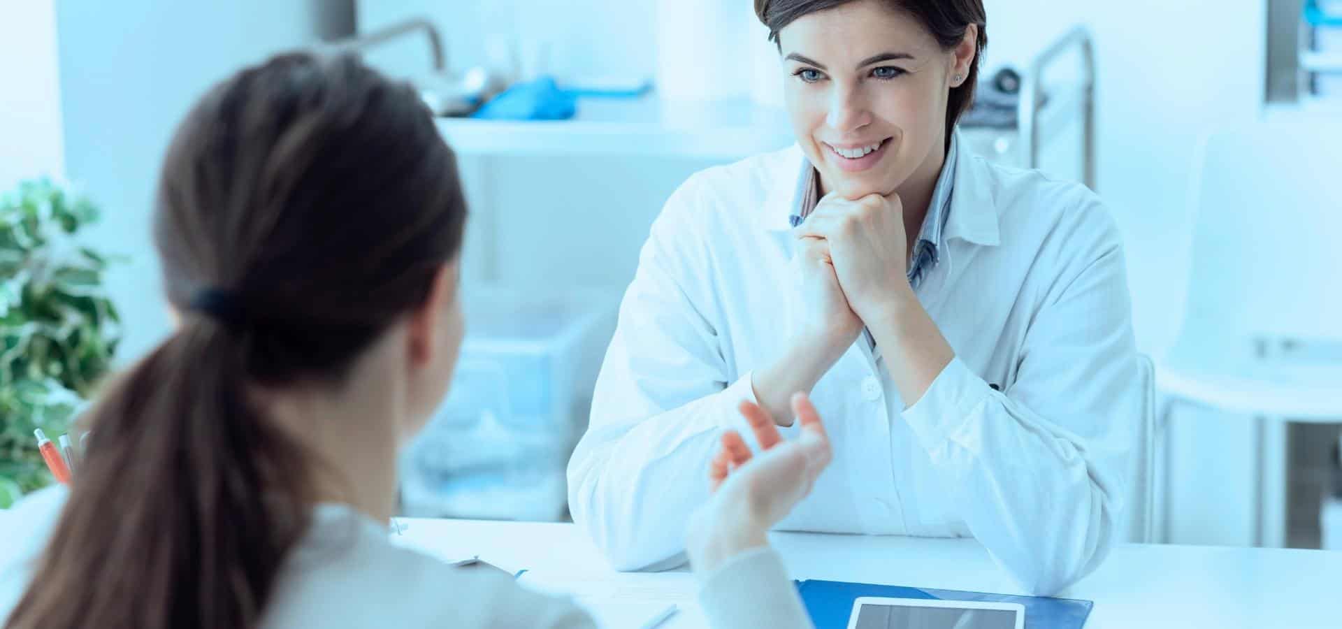 Eine Ärztin im weißen Kittel lächelt und hört einer Patientin während eines Gesprächs in einer hellen, modernen Klinik zu.