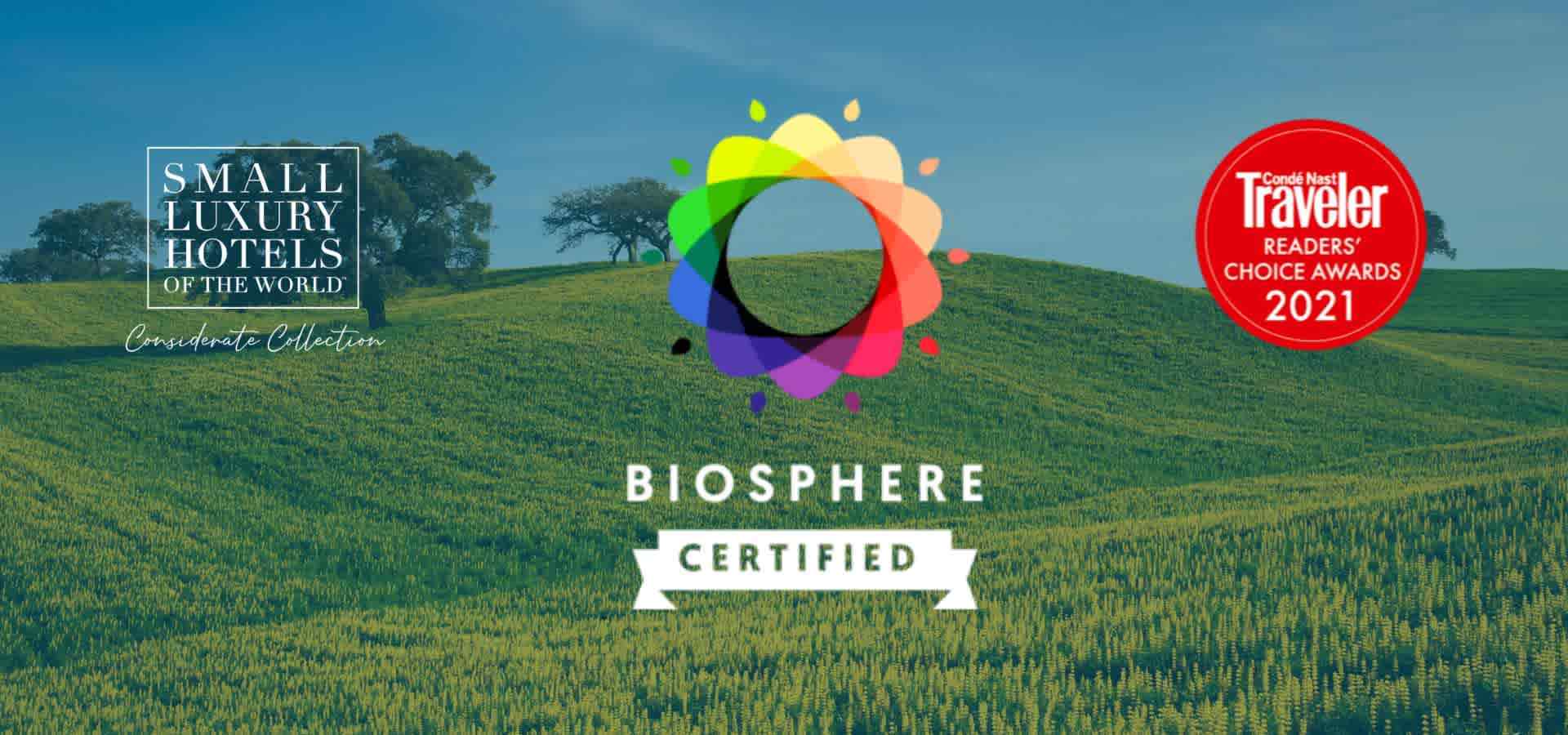 Awards and sustainability recognition graphic featuring hospitality and travel industry certifications displayed over a scenic countryside landscape with rolling green fields and scattered trees.