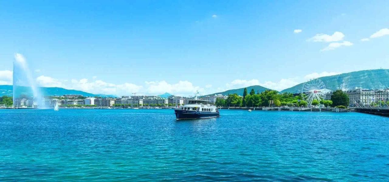 Panoramic view of a lakeside city with a passenger boat cruising across clear blue water, a towering fountain, a Ferris wheel along the waterfront, and rolling hills beneath a bright summer sky.