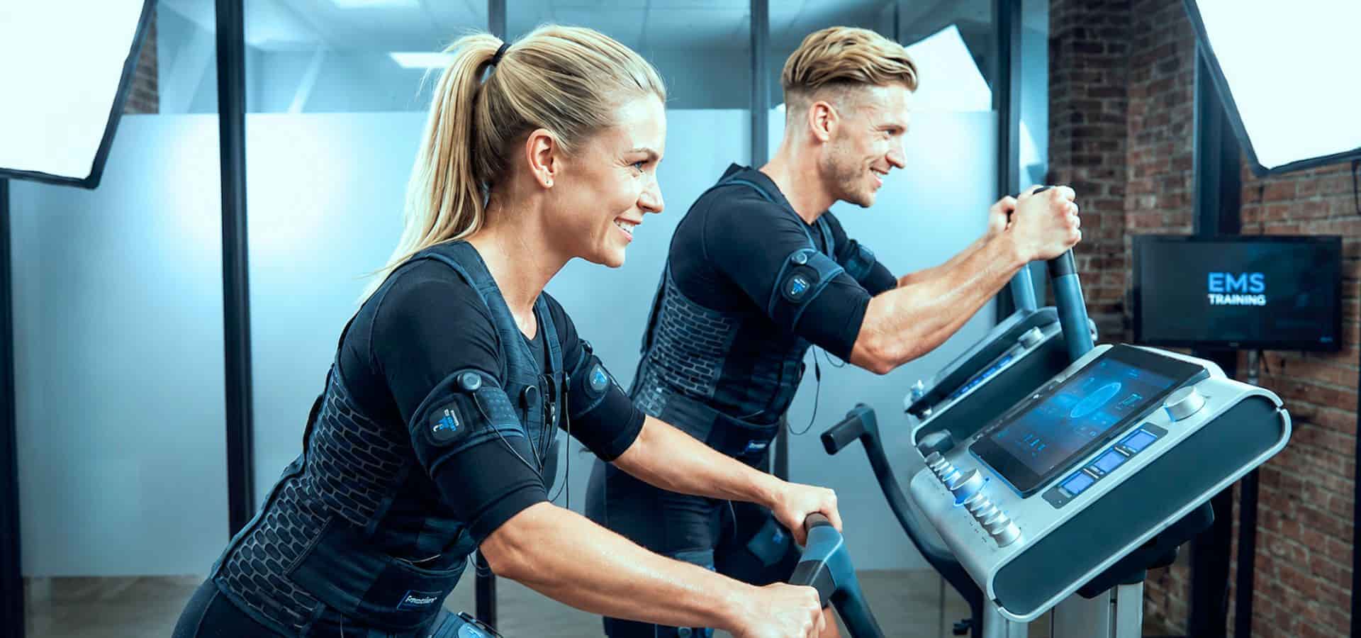 Two people wearing EMS training suits exercising on cardio machines in a modern fitness studio.