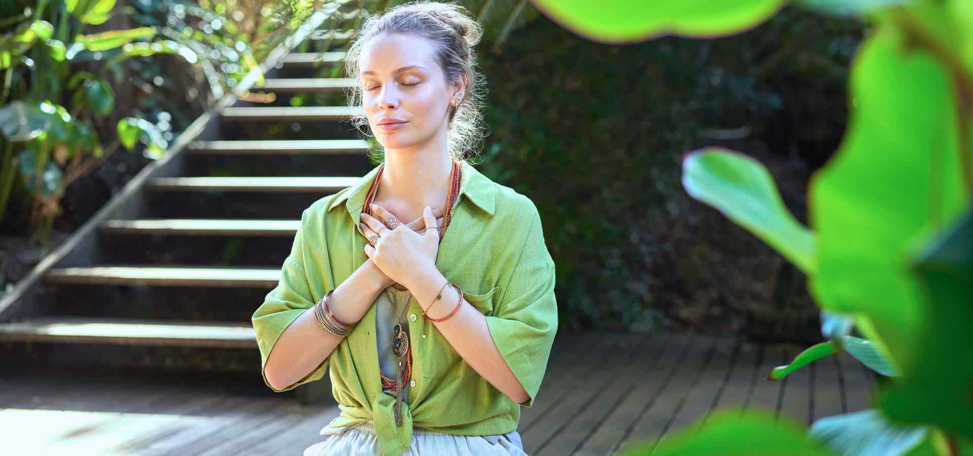 A woman practicing mindfulness meditation outdoors in a lush garden setting, sitting peacefully with her eyes closed and hands placed over her chest, surrounded by greenery and natural light.
