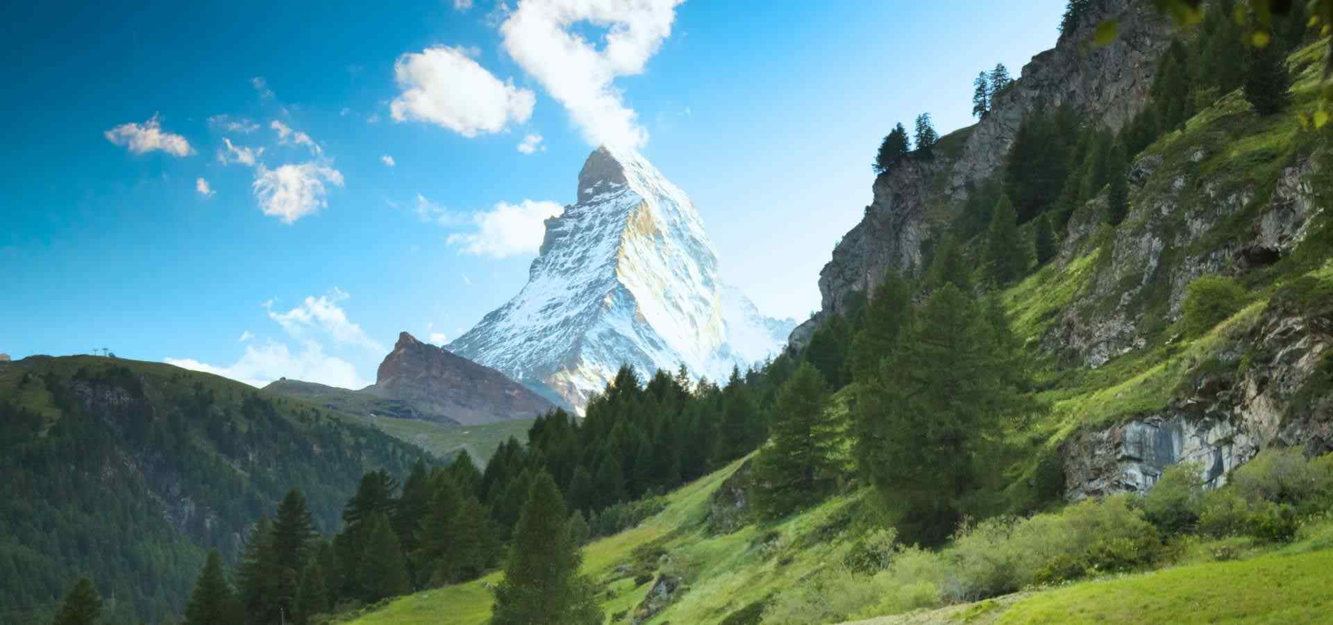 Dramatic view of a towering snow-capped alpine mountain peak rising above lush green valleys, pine forests, and rocky cliffs under a bright blue sky with scattered clouds.