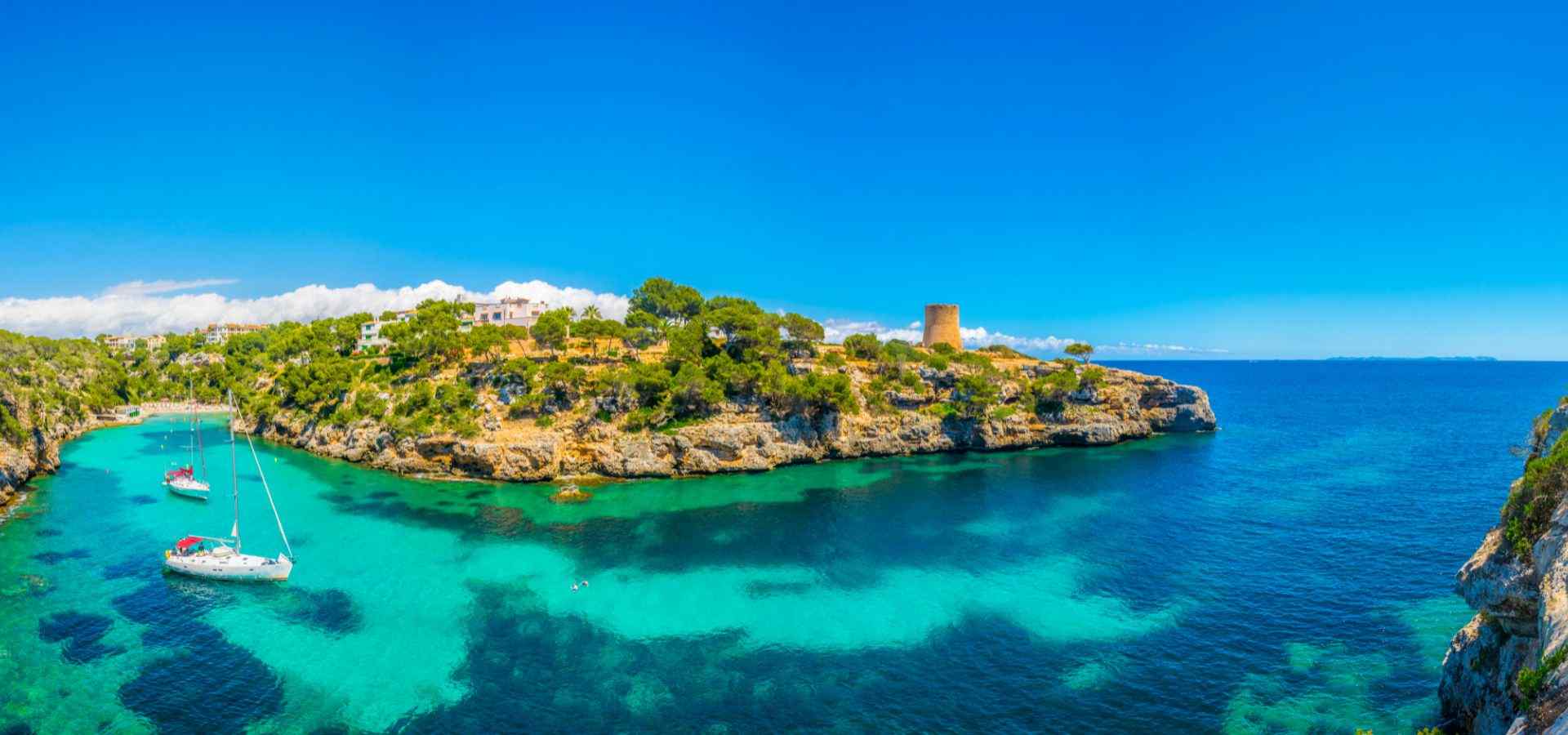 Panoramic view of a Mediterranean coastal bay with crystal-clear turquoise water, anchored sailboats, rocky cliffs, lush green vegetation, and a historic stone watchtower under a bright blue sky.
