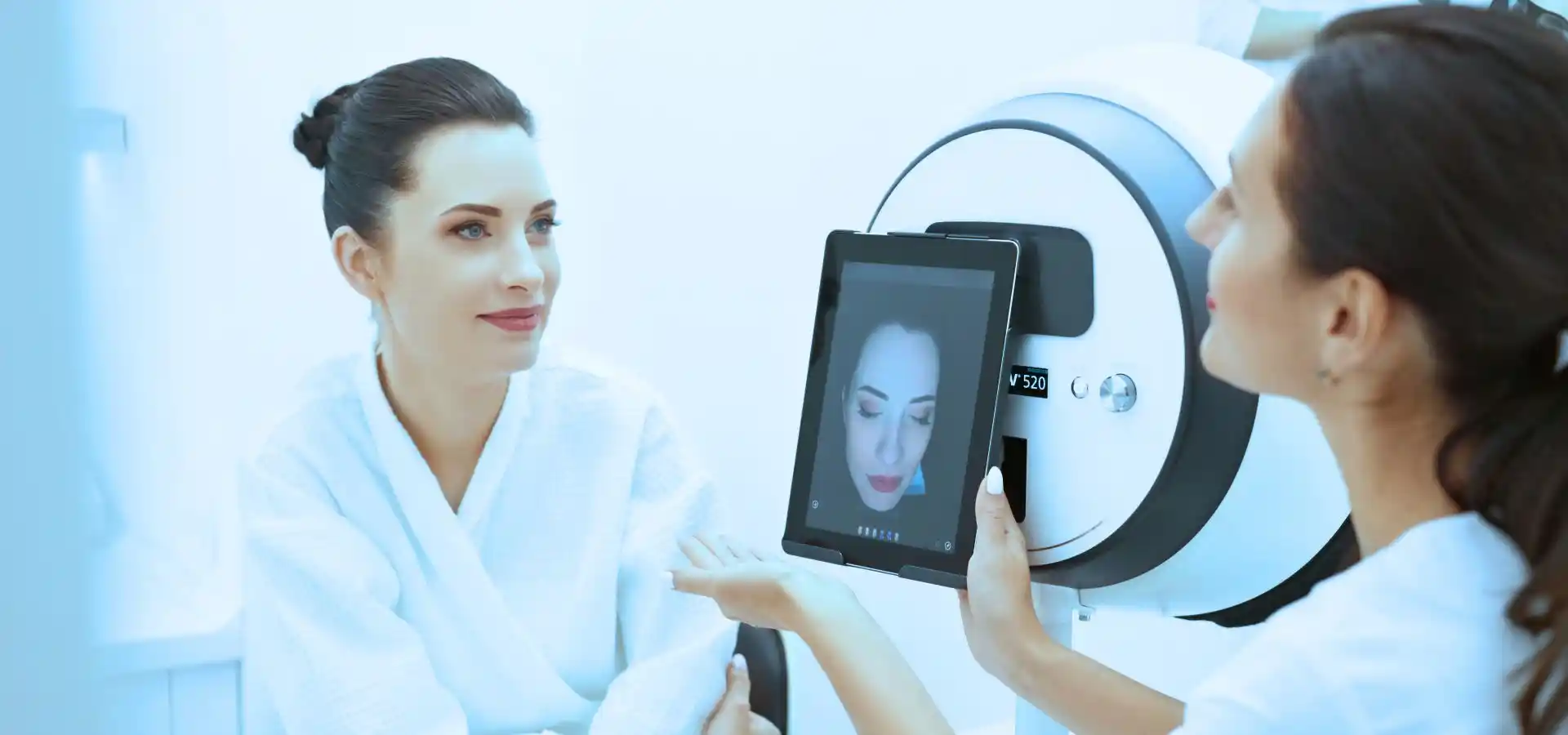 A woman in a white robe sits facing a skincare professional who is holding a device with a screen displaying the woman’s facial analysis in a clinical setting.