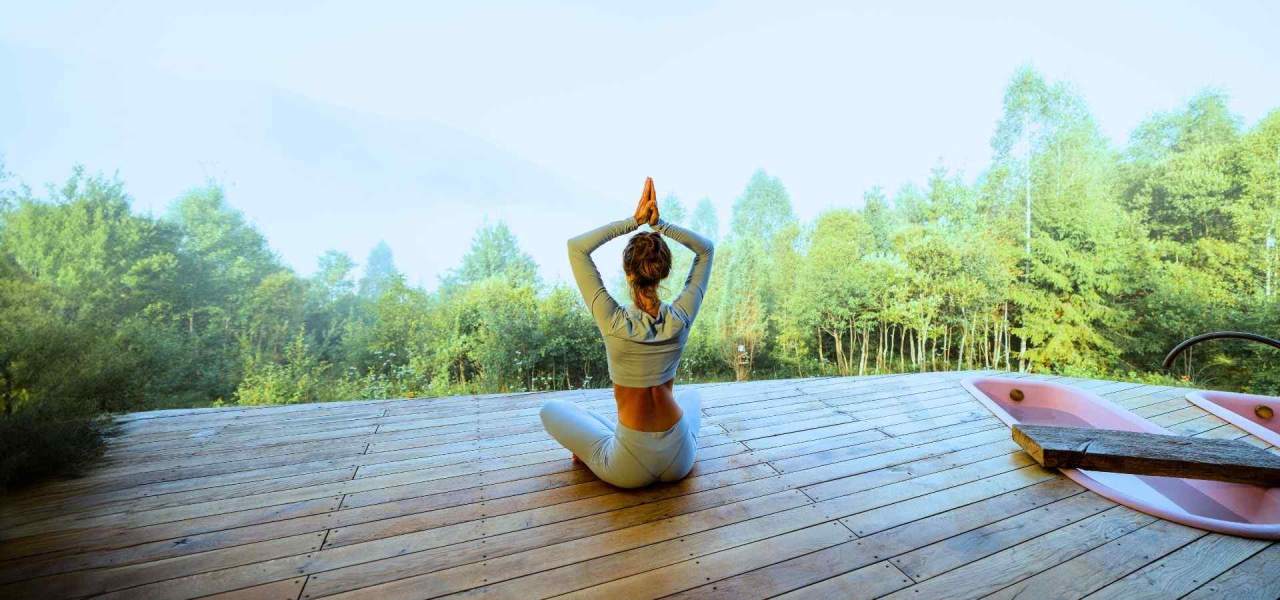 Woman practicing yoga meditation on a wooden wellness retreat deck surrounded by peaceful green forest and mountain scenery.
