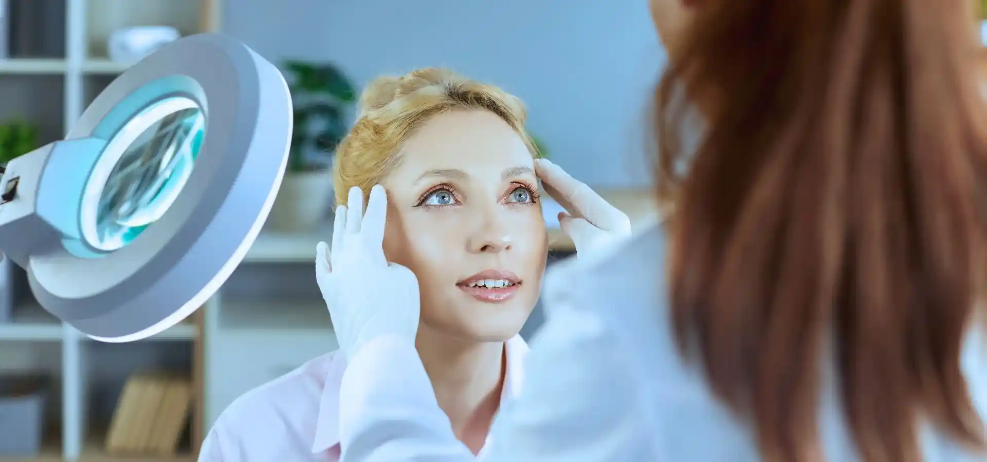 A skincare professional wearing white gloves examines a woman’s face, gently holding her temples, with a magnifying lamp nearby in a clinical setting.