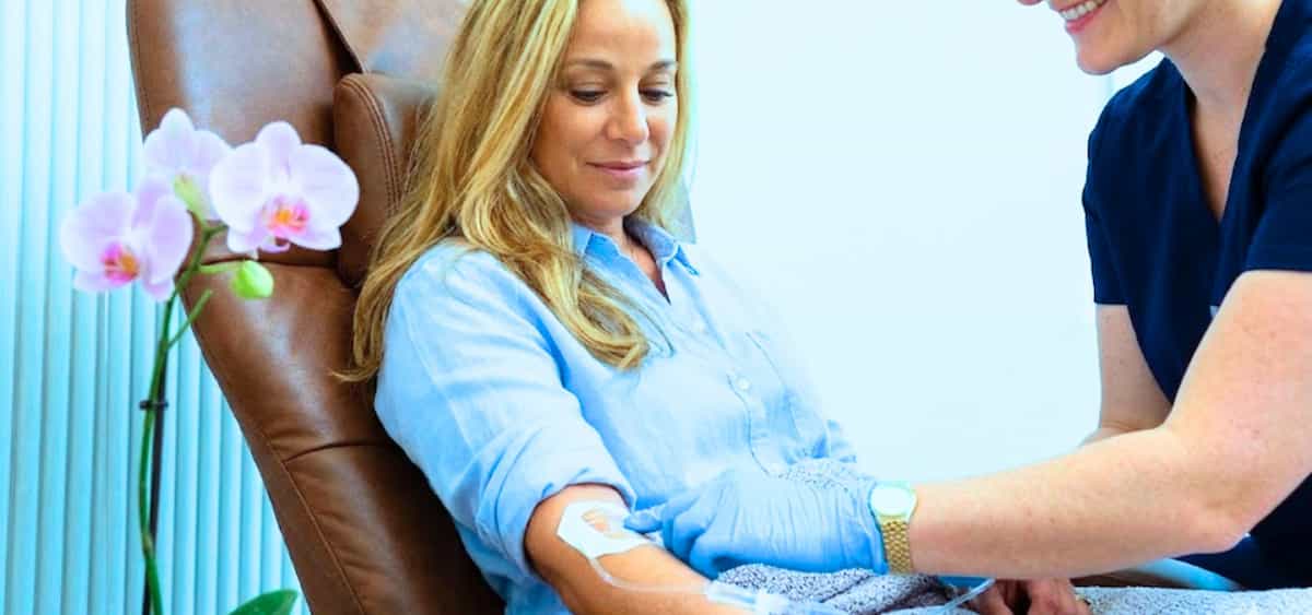 Woman sitting in a clinic chair receiving a medical test while a healthcare professional monitors her arm.