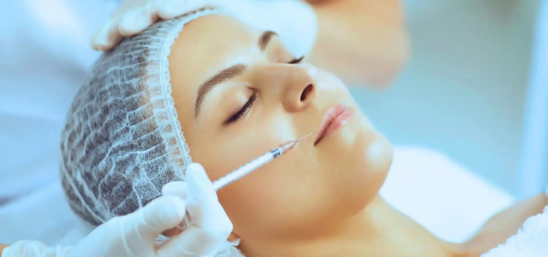 A woman lying on a treatment bed receiving a facial injection from a professional wearing gloves, with a hair cap on, in a clinical setting.