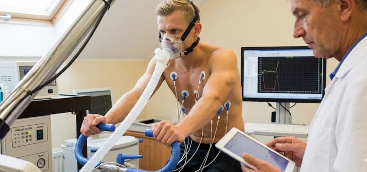 Man performing a medical fitness stress test on a stationary bike while wearing a breathing mask and chest sensors, as a doctor monitors results on a tablet and computer screen.
