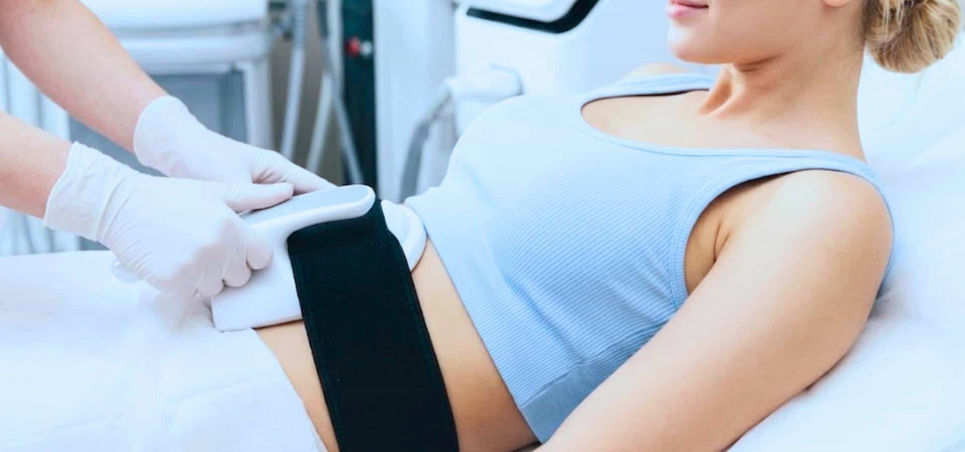 Patientin liegt auf einer Behandlungsliege, während ein medizinischer Fachmann ein Handgerät am Bauch für eine klinische Behandlung verwendet.