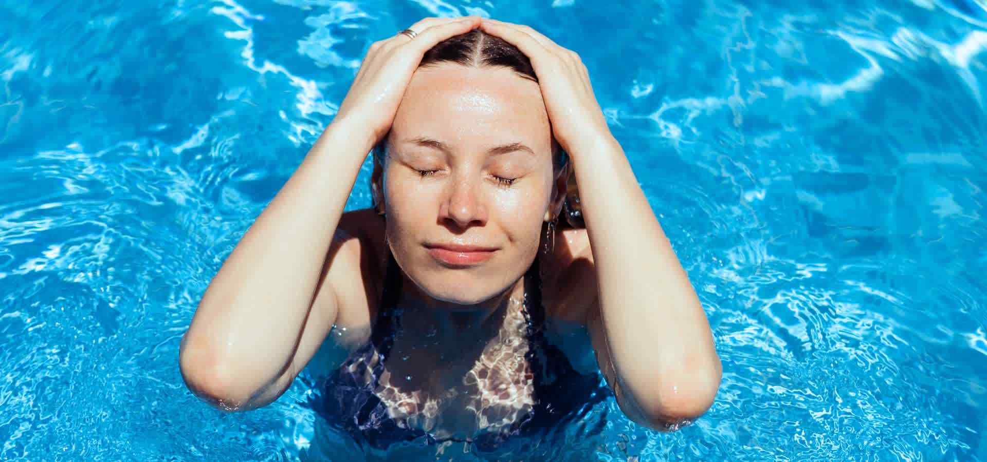 A woman relaxing in a swimming pool with her eyes closed, gently holding her head above crystal-clear blue water while enjoying a peaceful wellness and hydrotherapy experience.