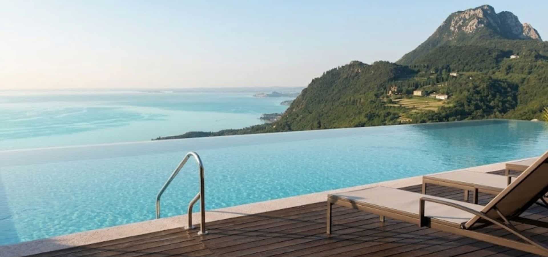 Infinity pool overlooking Lake Garda with lounge chairs, calm blue water, and green mountains under a clear sky.