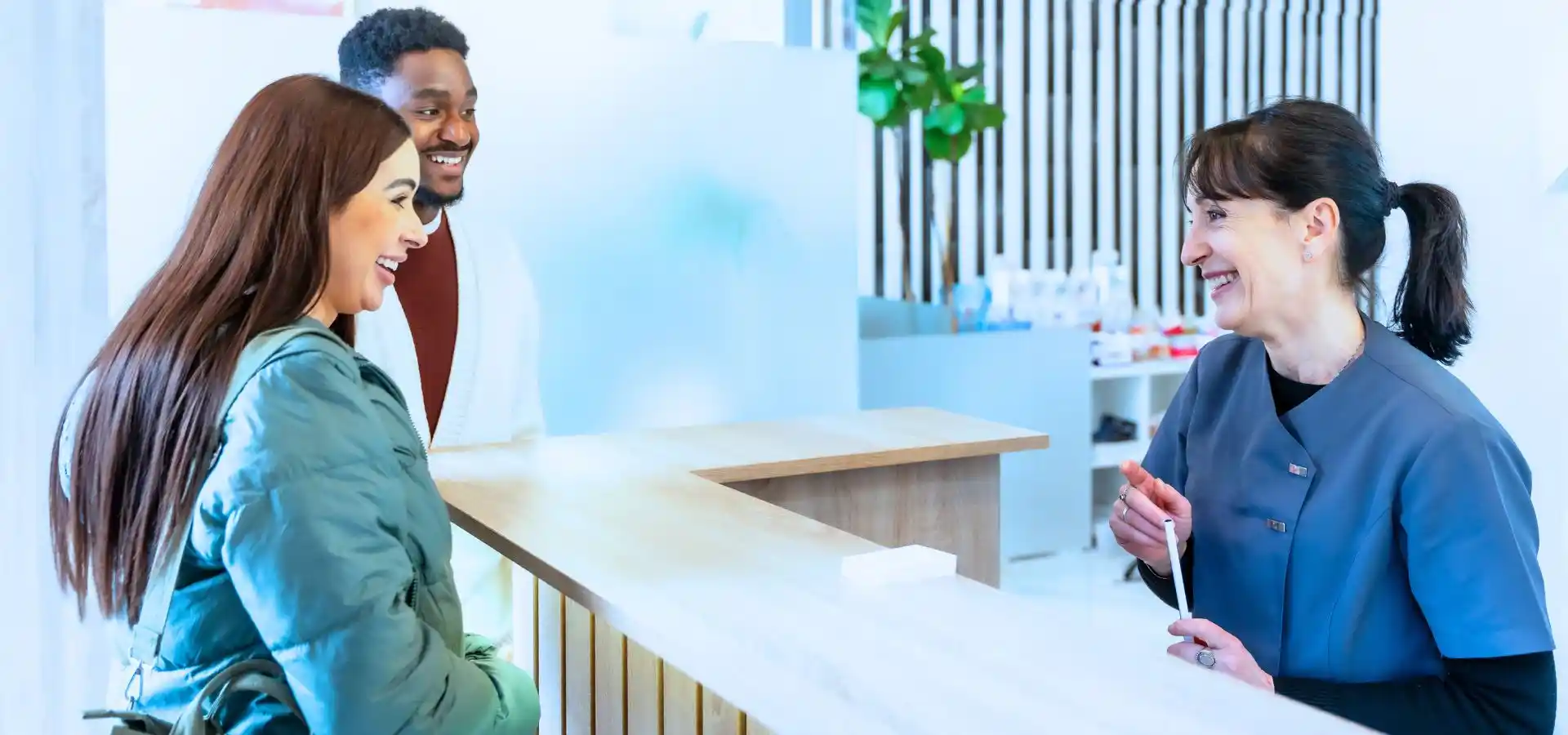 Patients speak with a clinic receptionist at a modern medical reception desk in a bright healthcare facility.