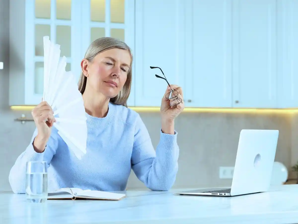 Middle-aged woman experiencing hot flashes while fanning herself at home, with a laptop, notebook, and glass of water nearby.