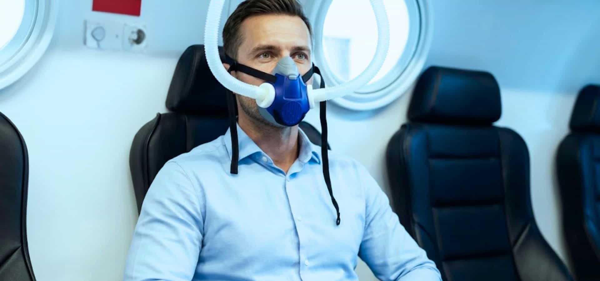 Man sitting in a medical chair wearing a breathing mask connected to testing equipment, undergoing a respiratory or metabolic assessment in a clinical setting.