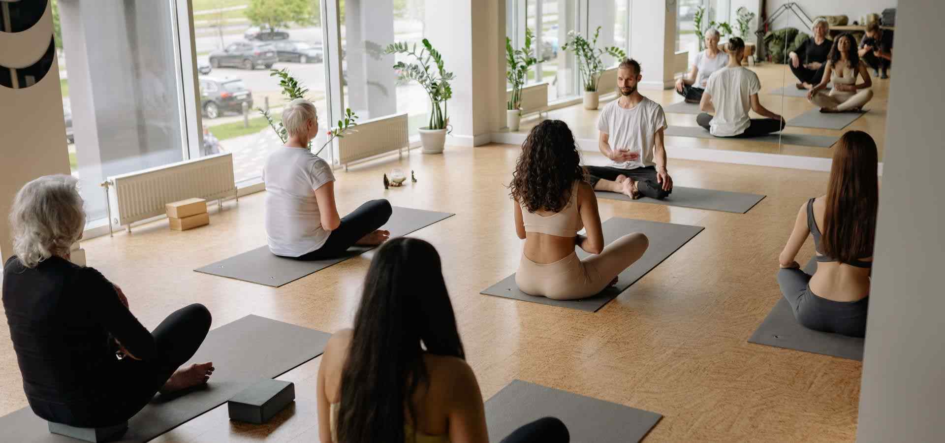 Group meditation and yoga session in a bright wellness studio, with participants seated on yoga mats while listening to an instructor, surrounded by natural light, mirrors, and indoor plants.