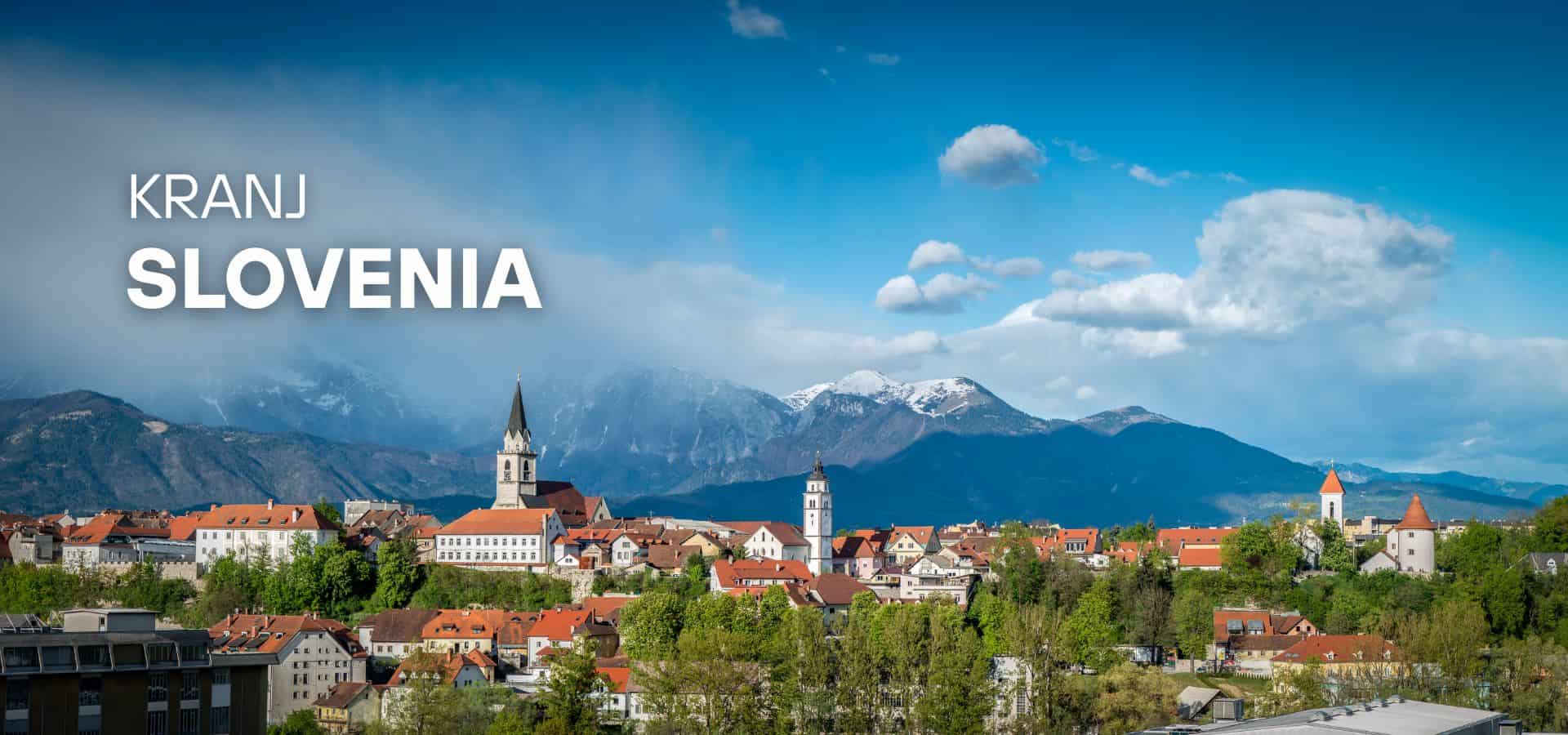 Panoramic view of the town of Kranj, Slovenia, featuring red-roofed buildings and historic church steeples against a backdrop of the snow-capped Kamnik-Savinja Alps under a blue sky.