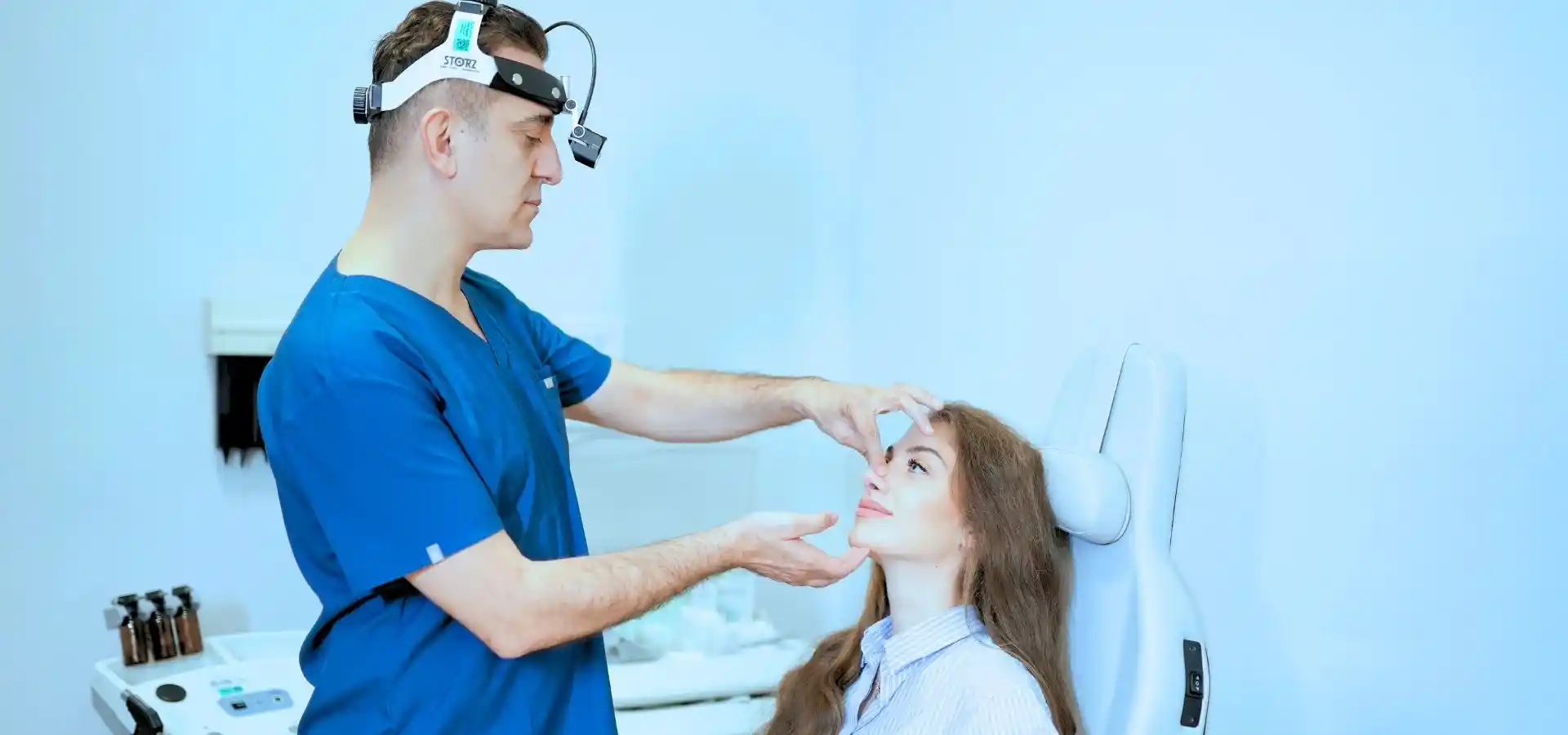 A healthcare professional wearing a head-mounted examination light performs a facial assessment on a seated patient in a modern medical clinic.