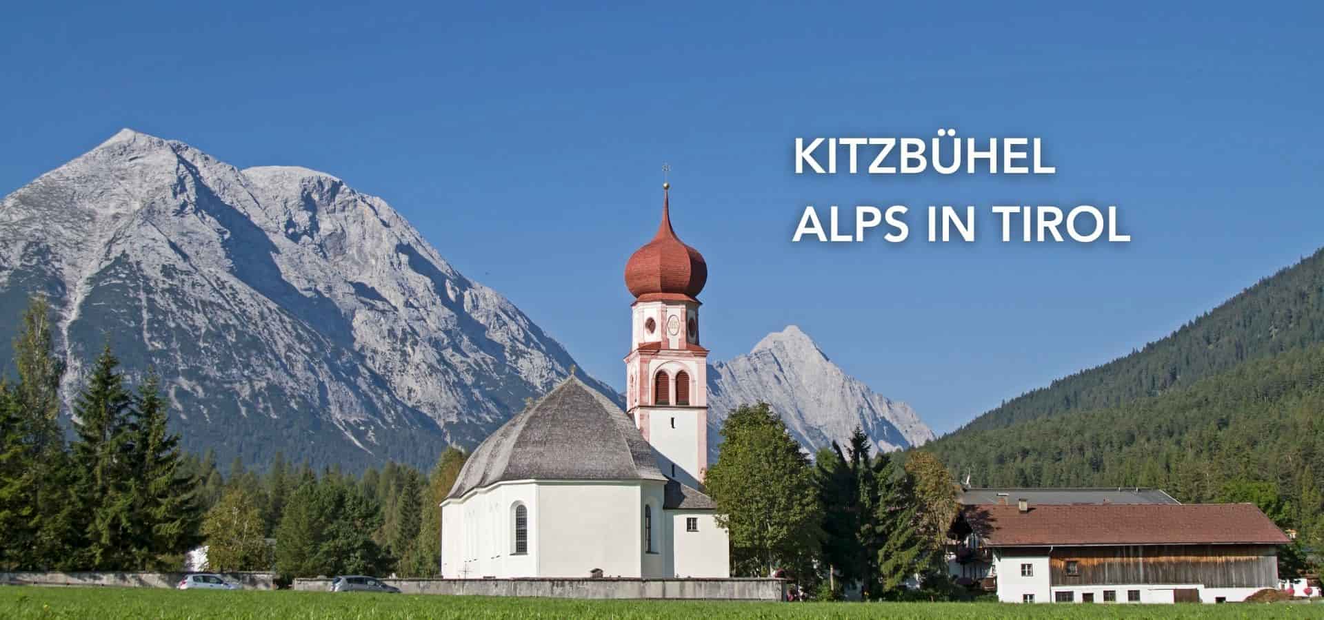 Scenic view of Kitzbühel in the Tirol Alps featuring a traditional church, alpine houses, green forests, and towering mountains under a clear blue sky.
