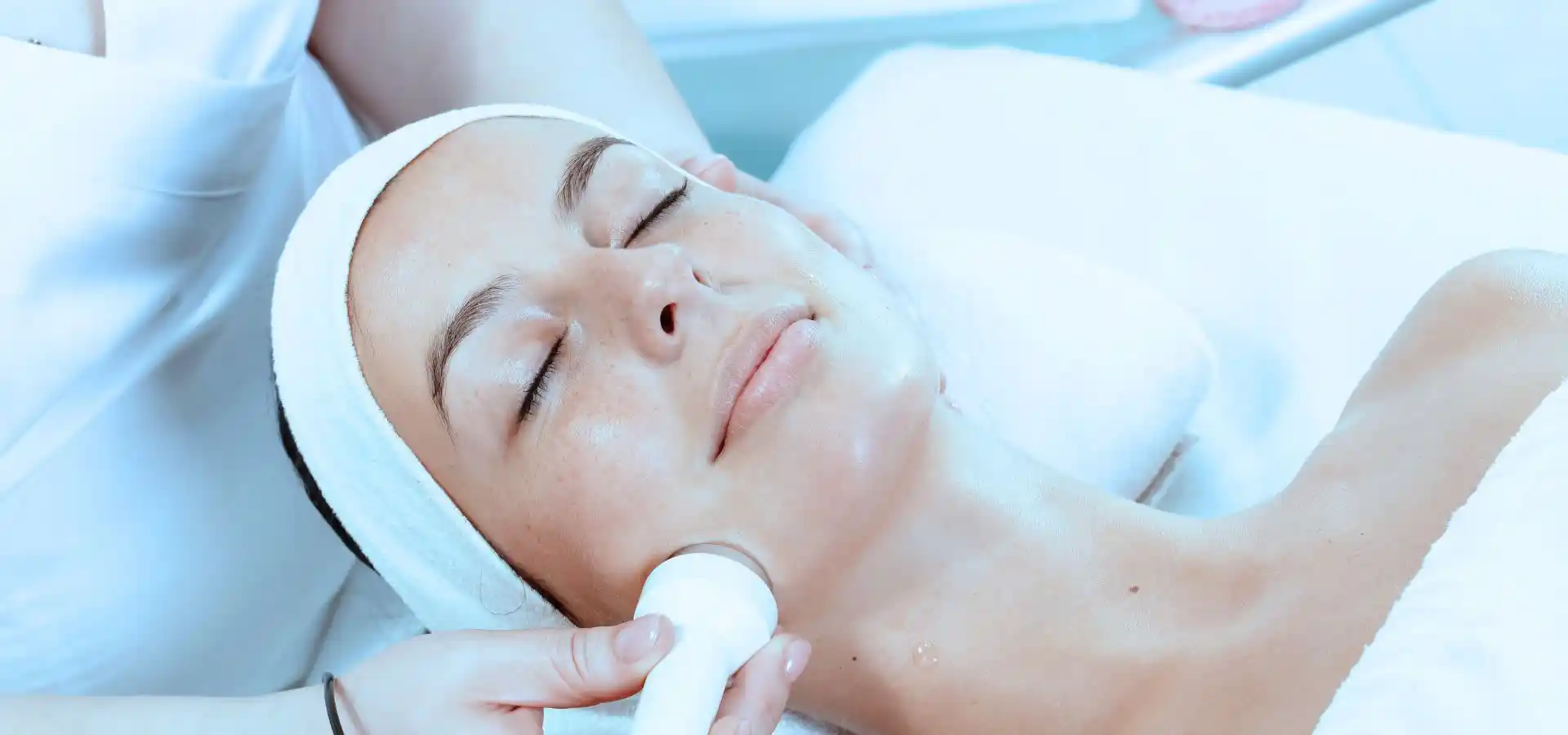 A person relaxes with their eyes closed while receiving a professional facial treatment, as a skincare specialist uses a handheld device on the jawline in a modern aesthetic clinic.
