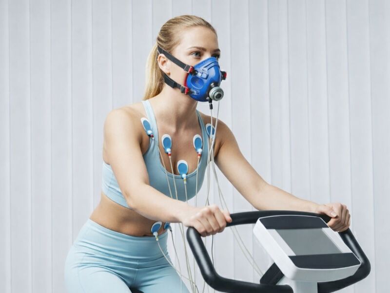 Woman wearing a respiratory mask and chest electrodes while cycling on a stationary bike during a fitness performance test.