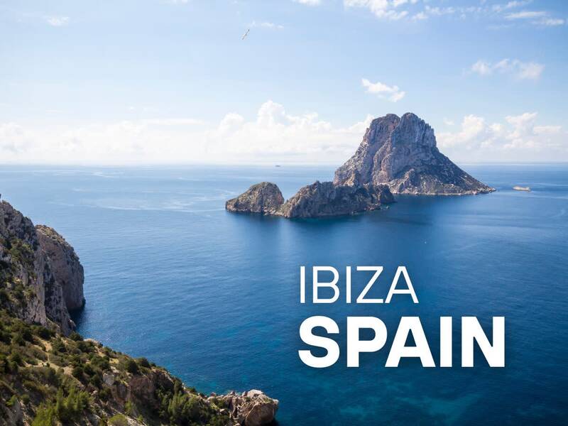 Coastal view of the iconic rocky island of Es Vedrà in Ibiza, Spain. The image features deep blue water, rugged cliffs in the foreground, and the words 'IBIZA SPAIN' overlaid in white text.